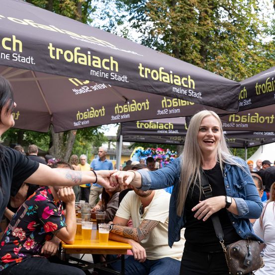 Eine Frau mit langen blonden Haaren lächelt und schüttelt einem Mann die Hand bei einer Veranstaltung im Freien. Sie sind von Menschen umgeben, die an Tischen mit Getränken sitzen. Der Baldachin darüber hat gelben Text.