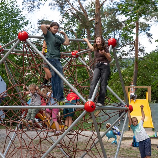 Mehrere Kinder spielen auf einer großen Metall-Kletterstruktur in einem Park. Zwei Mädchen stehen oben, während andere um sie herum klettern.