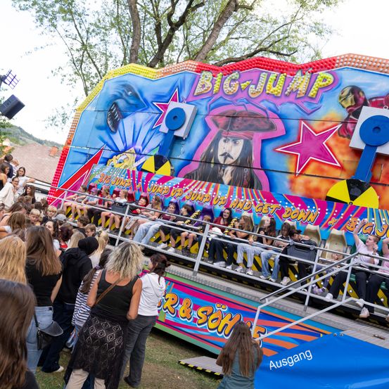 Eine Menge Menschen versammelt sich um eine bunte Karussellfahrt namens 'Big Jump' mit einem Piratencharakter an der Seite. Einige warten darauf zu fahren, während andere zuschauen.