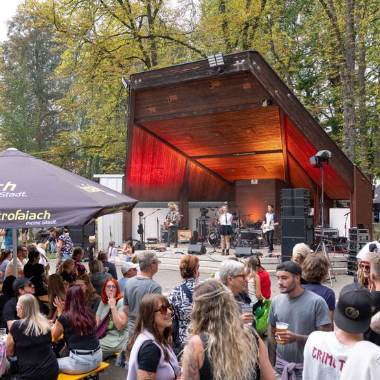 Eine Band spielt auf einer Bühne in einer Outdoor-Umgebung, mit einer großen Menschenmenge, die sich versammelt hat. Die Bühne ist mit roter Beleuchtung bedeckt und das Publikum sitzt auf Bänken und steht.