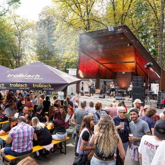 Ein lebhaftes Open-Air-Event mit einer Menge, die Getränke genießt und Musik unter Schirmen hört. Eine Bühne mit Musikern im Hintergrund.