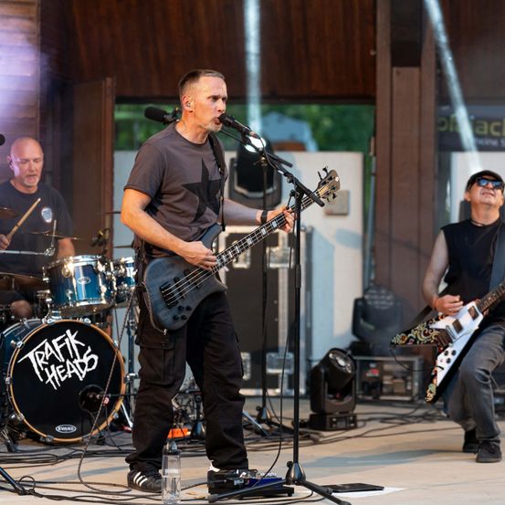 Eine Band spielt auf der Bühne, ein Gitarrist singt in ein Mikrofon, ein Schlagzeuger hinter ihm und ein weiterer Gitarrist auf der rechten Seite. Das Schlagzeug des Schlagzeugers hat das Wort 'Trafik' darauf.