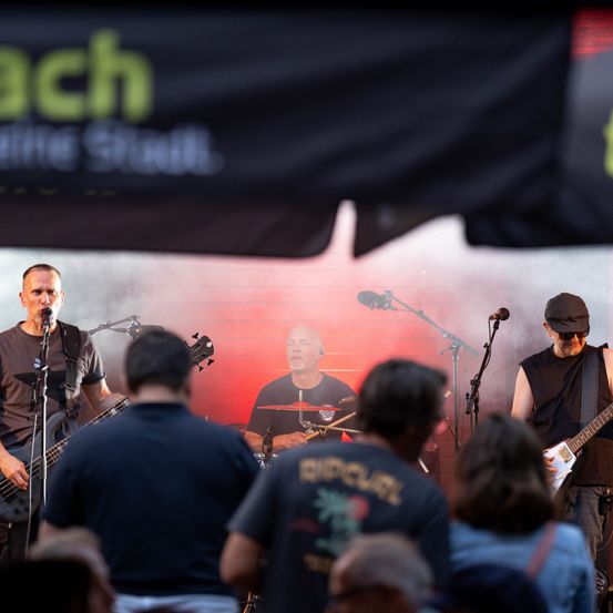 Eine Band tritt auf der Bühne auf, mit einem Banner im Hintergrund, das 'Beach' steht. Der Leadsänger singt in ein Mikrofon, während der Gitarrist und der Schlagzeuger ihre Instrumente spielen. Das Publikum schaut von vorne zu.
