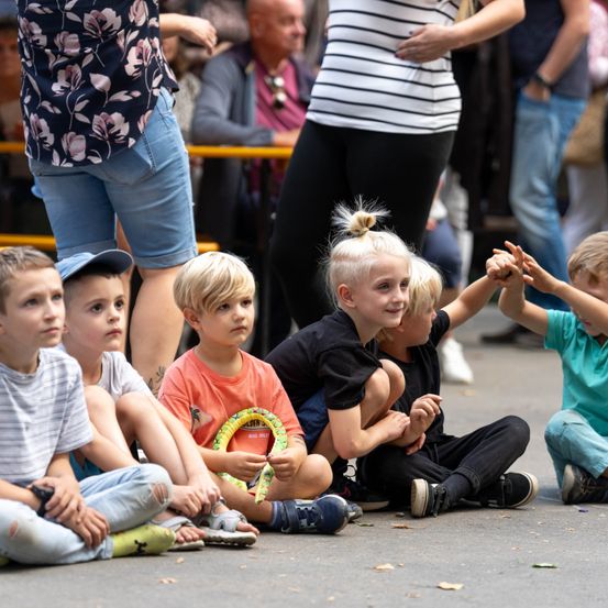 Eine Gruppe junger Jungen sitzt auf dem Boden, einige halten Händchen, mit Erwachsenen im unscharfen Hintergrund.
