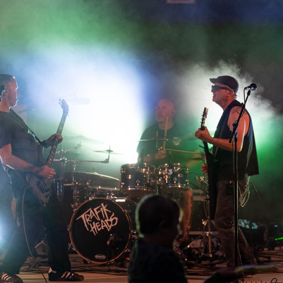 Eine Rockband tritt auf der Bühne auf, mit grünem und weißem Licht. Der Leadgitarrist spielt eine E-Gitarre, während der Schlagzeuger hinter einem Schlagzeug spielt. Der Name der Band, 'Traffic Heads', steht auf dem Schlagzeug.