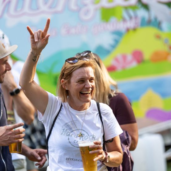 Eine Frau in einem weißen T-Shirt mit Friedenszeichen und einem Bier in der Hand, lächelt und ist von anderen bei einer Veranstaltung im Freien umgeben.