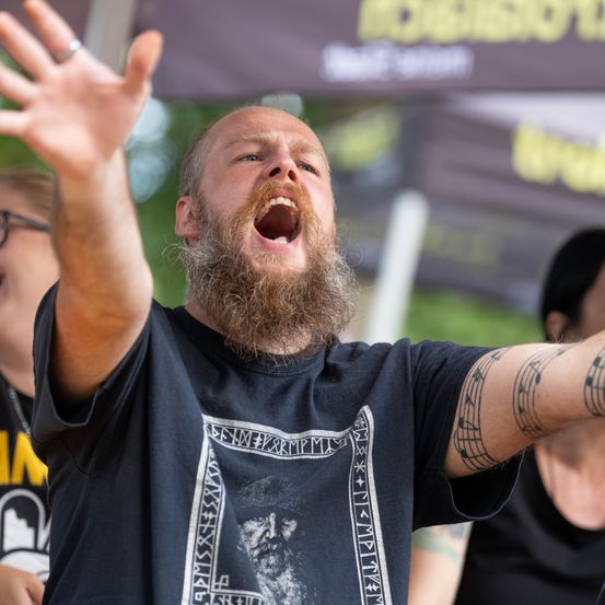Ein Mann mit Bart und Tattoos schreit mit erhobenen Armen bei einer Veranstaltung im Freien.
