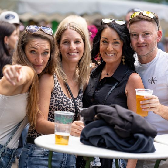 Vier Menschen lächeln und posieren für ein Foto bei einer Outdoor-Veranstaltung. Zwei Frauen und zwei Männer, ein Mann hält ein Bierglas.