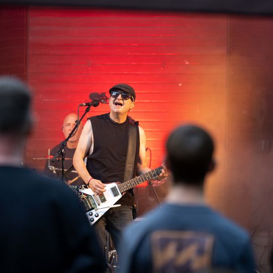 Ein Mann mit Hut und Sonnenbrille spielt eine E-Gitarre auf der Bühne. Hinter ihm spielt ein anderer Mann Schlagzeug. Das Publikum schaut zu.