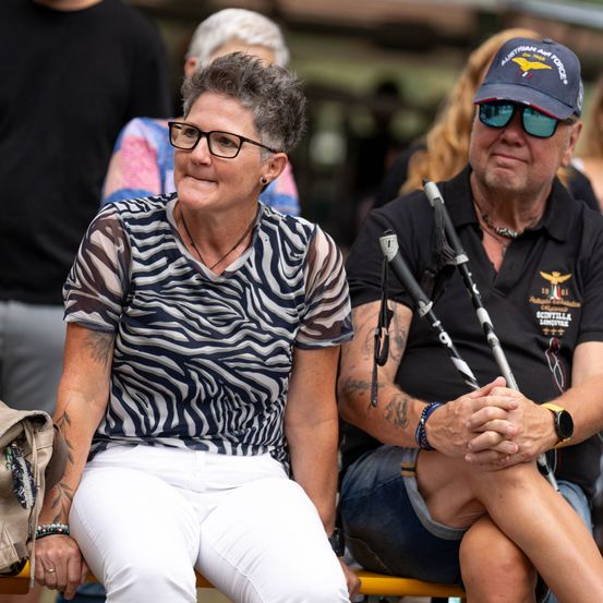 Eine Frau mit grauem Haar trägt ein Zebra-Muster-Shirt und weiße Hosen. Sie sitzt neben einem Mann in einem schwarzen Shirt mit einer Kappe und Sonnenbrille. Sie schauen beide nach vorne.