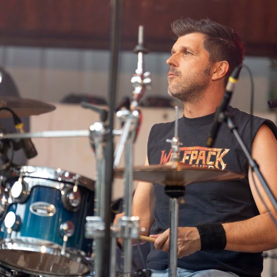 Ein Mann mit einem schwarzen ärmellosen Shirt mit dem Wort 'Wetpack' spielt die Drums auf der Bühne.