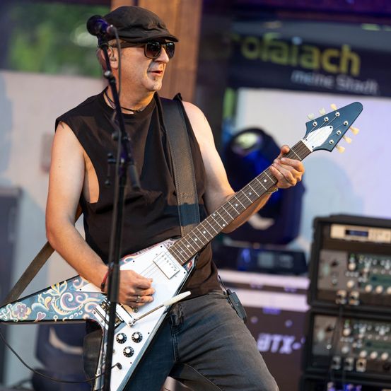 Ein Mann in einem schwarzen, ärmellosen Shirt und Jeans spielt Gitarre auf der Bühne. Er trägt eine Sonnenbrille und einen Hut.