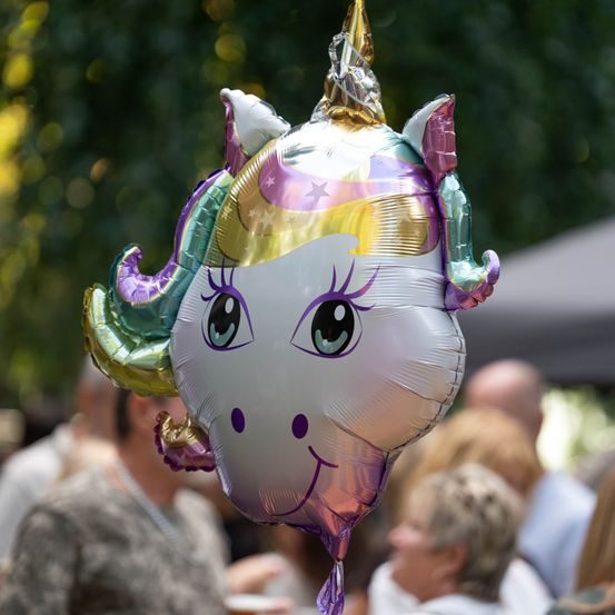 Ein Nahaufnahme eines farbenfrohen Einhorn-Ballons mit goldenem Horn und lächelndem Gesicht, der bei einer Veranstaltung im Freien in der Luft schwebt.
