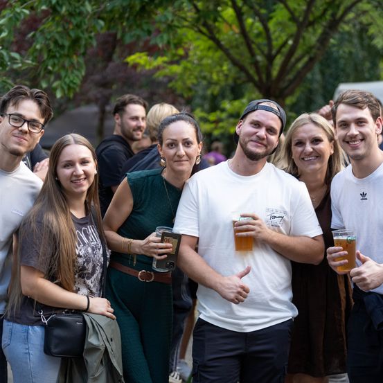 Eine Gruppe von Freunden in lässiger Kleidung, die lächeln und Gläser mit Bier in der Hand halten, stehen in einer Außenumgebung mit Bäumen im Hintergrund.