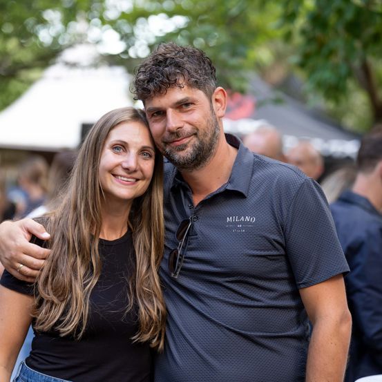 Ein lächelndes Paar steht eng beieinander, der Mann trägt ein Milan-Shirt und die Frau ein schwarzes Shirt, und posiert für ein Foto bei einer Outdoor-Veranstaltung.