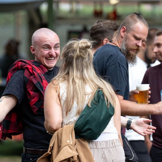 Eine Gruppe von Menschen bei einer Outdoor-Veranstaltung, ein Mann mit Bart hält ein Bierglas, ein anderer Mann lächelt und eine Frau mit Dutt.