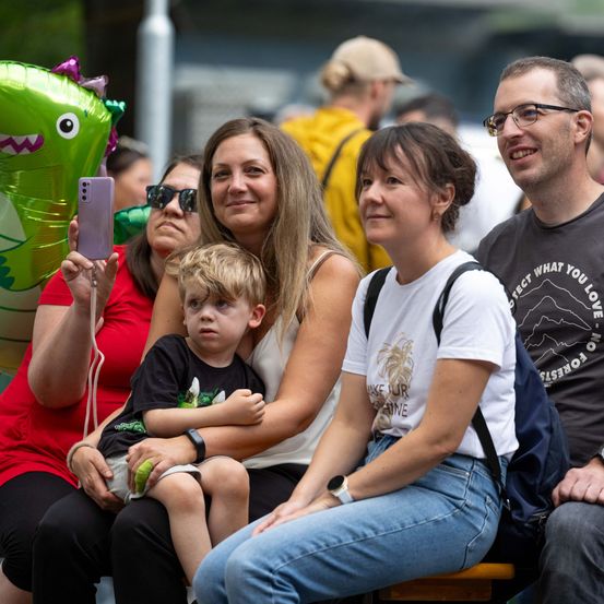 Eine Gruppe von Menschen, darunter eine Frau, die ein lila Telefon hält, ein Kind und zwei Frauen, sitzen zusammen und lächeln. Sie sind bei einer Veranstaltung im Freien, mit einem grünen Dinosaurierballon und einem Mann mit Brille im Hintergrund.