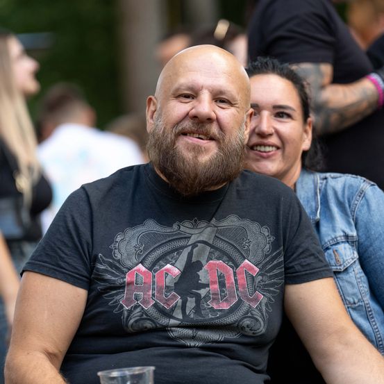 Ein Mann mit Bart trägt ein AC/DC-T-Shirt und sitzt neben einer Frau bei einer Outdoor-Veranstaltung.