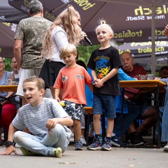 Drei Kinder stehen bei einer Outdoor-Veranstaltung zusammen, eine Frau mit einem Mikrofon steht hinter ihnen. Die Kinder lächeln und schauen in die Kamera.