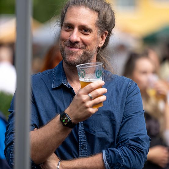 Ein Mann mit Bart und langen Haaren, in einem blauen Hemd, hält einen Becher Bier mit einem Lächeln. Im Hintergrund sind verschwommen andere Leute bei einer Outdoor-Veranstaltung zu sehen.