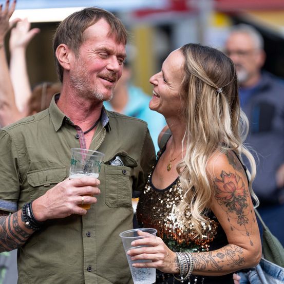 Ein Mann und eine Frau auf einem Festival, lächelnd und Getränke in der Hand. Der Mann hat Tattoos an den Armen, die Frau hat ein Blumentattoo am Arm.