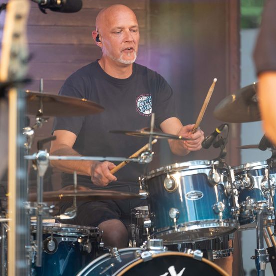 Ein kahlköpfiger Mann in einem schwarzen T-Shirt spielt die Drums bei einer Outdoor-Veranstaltung. Er hält Drumsticks und trägt Kopfhörer.