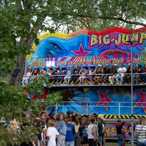 Ein farbenfroher 'Big Jump'-Karussell mit Menschen auf den Sitzen. Das Karussell ist von einer Menschenmenge umgeben. Bäume und ein Lautsprecher sind im Hintergrund zu sehen.