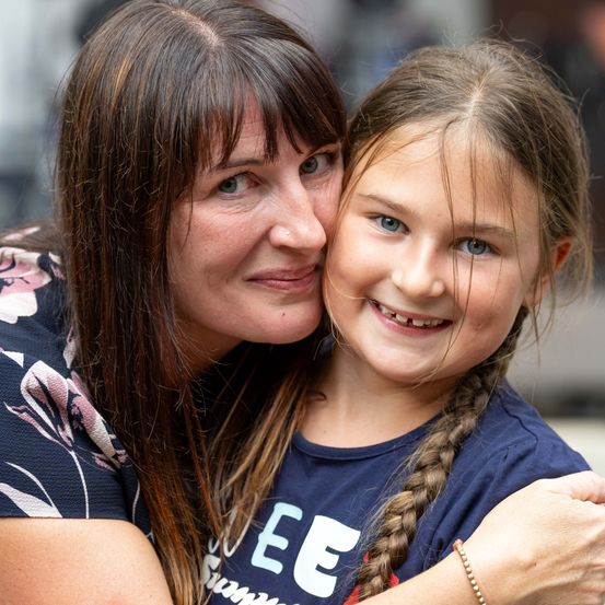 Eine lächelnde Frau mit braunen Haaren umarmt ein junges Mädchen mit geflochtenen Haaren und blauen Augen. Das Mädchen trägt ein blaues T-Shirt mit weißen Buchstaben.