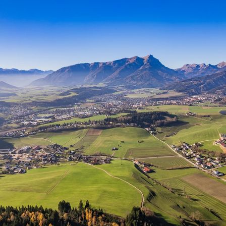 Luftaufnahme eines Tals mit grünen Feldern, einem kleinen Städtchen und Bergen in der Ferne unter einem klaren blauen Himmel.