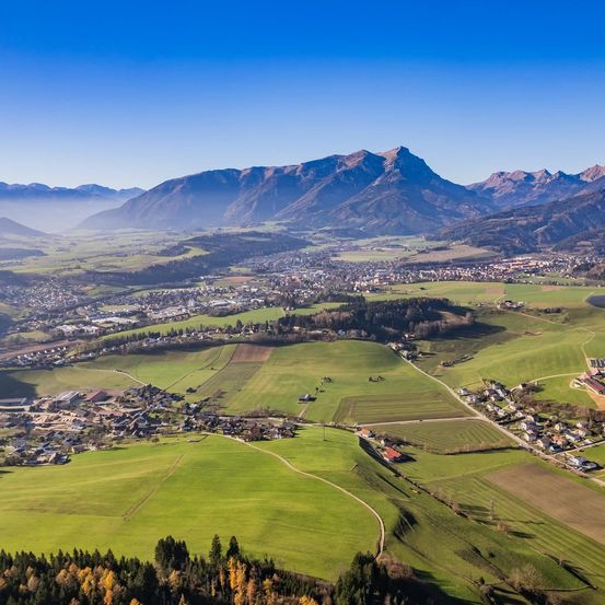 Luftaufnahme eines Tals mit grünen Feldern, einem kleinen Städtchen und Bergen in der Ferne unter einem klaren blauen Himmel.