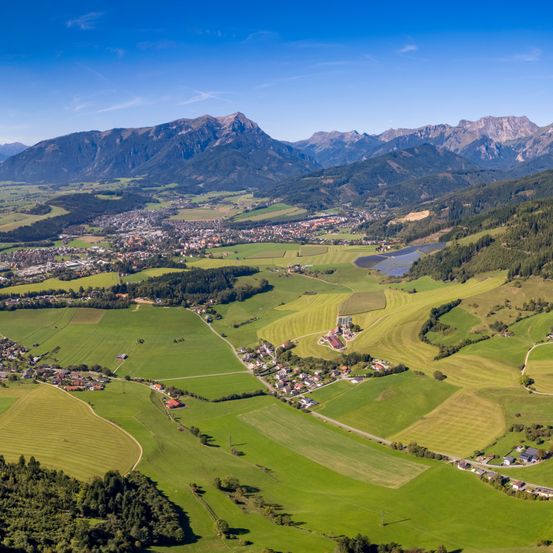 Luftaufnahme eines Dorfes, umgeben von üppigen grünen Feldern und Bergen unter einem klaren blauen Himmel.