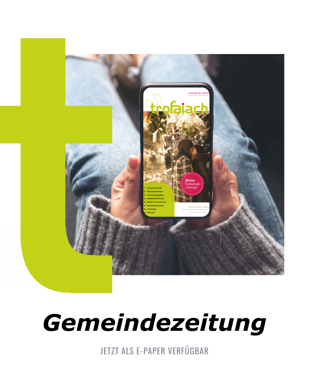 Eine Person hält ein Handy mit der Anzeige der 'Trofaiach' Gemeindezeitung. Das Cover zeigt ein Paar unter Weihnachtslichtern. Darunter ist ein grüner 'Gemeindzeitung' Text zu sehen.