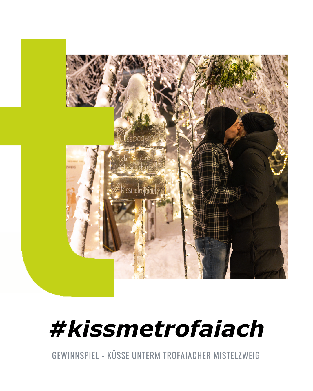 Ein Paar küsst sich unter einem schneebedeckten Bogen mit Weihnachtslichtern in einer winterlichen Umgebung. Der Schild sagt 'Kissbogen'. Der Hashtag #kissmetrofaiach ist sichtbar.