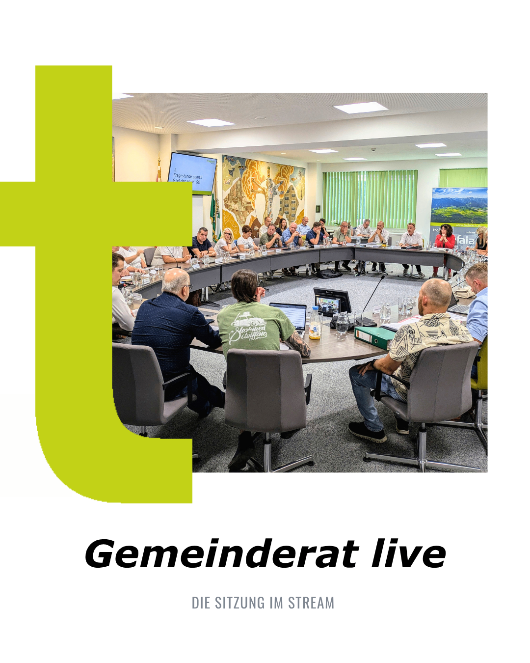 Eine laufende Ratssitzung, bei der Ratsmitglieder um einen Tisch herum sitzen, ein Monitor an der Wand und Personen im Hintergrund stehen.