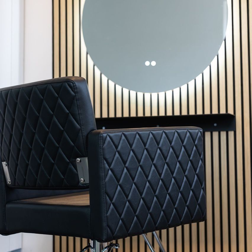 Ein schwarzer Lederdrehstuhl mit Diamantmuster steht vor einer Holzwand mit einem großen grauen runden Spiegel.