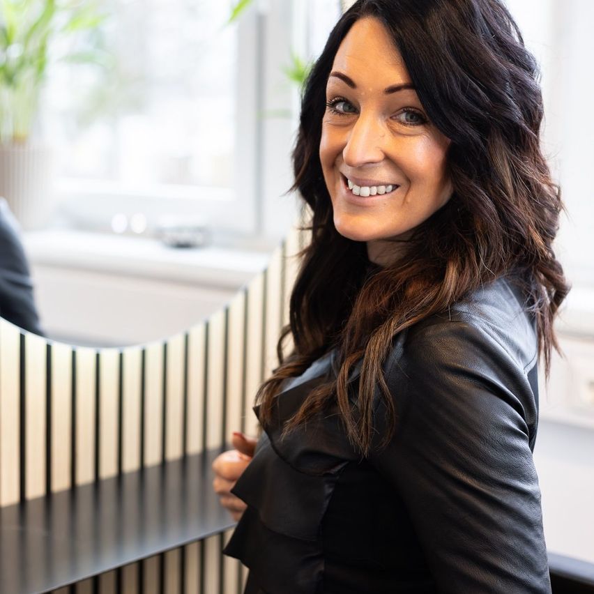 Eine lächelnde Frau mit langen, welligen braunen Haaren und einer schwarzen Lederjacke steht vor einem Fenster. Dahinter sind eine verschwommene Figur und ein Topf mit Pflanzen zu sehen.