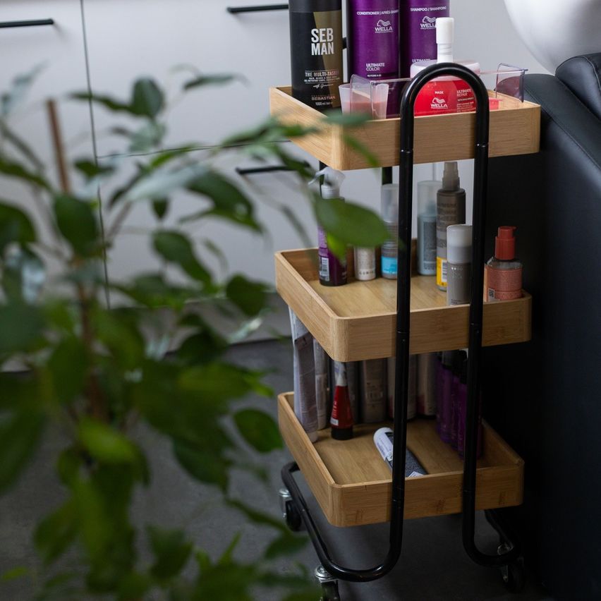Ein dreistufiger Bambuswagen hält verschiedene Haarpflegeprodukte, einschließlich einer roten Flasche, neben einer Pflanze und einem schwarzen Ledersofa.