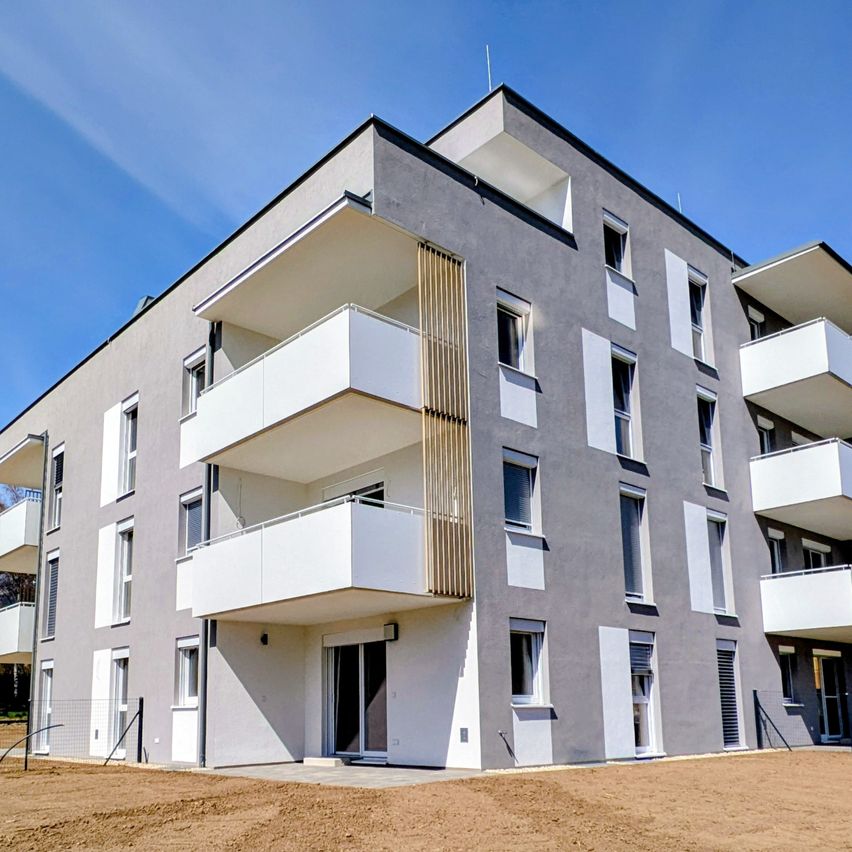 Ein modernes Apartmentgebäude mit mehreren Balkonen und Glastüren. Das Äußere ist grau mit weißen Akzenten, und die Balkone haben Holzgeländer. Das Gebäude steht vor einem klaren blauen Himmel.