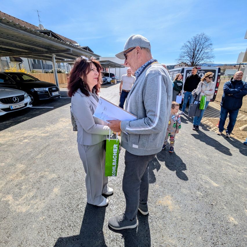 Ein älteres Paar steht auf einem Parkplatz, hält ein Dokument in der Hand, mit anderen dahinter und Autos unter einem Carport geparkt.
