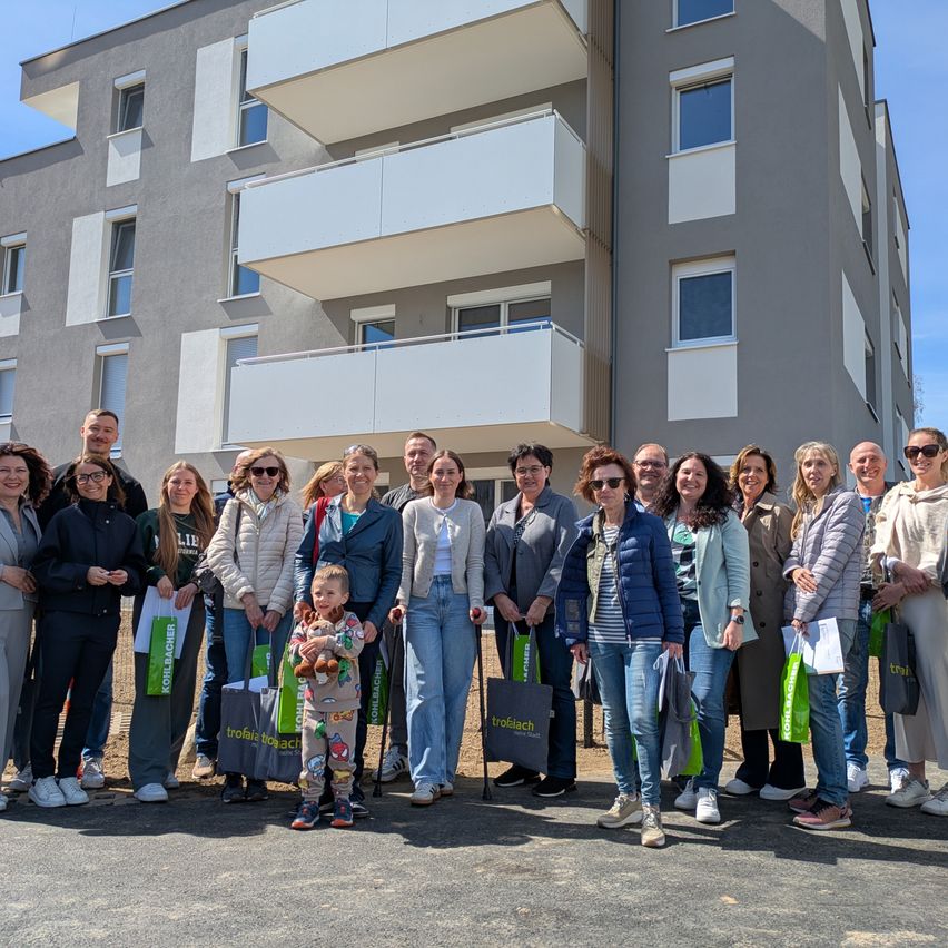 Eine Gruppe von Menschen steht vor einem Wohngebäude, trägt Freizeitkleidung und hält Einkaufstüten. Sie lächeln und scheinen für ein Foto zu posieren.