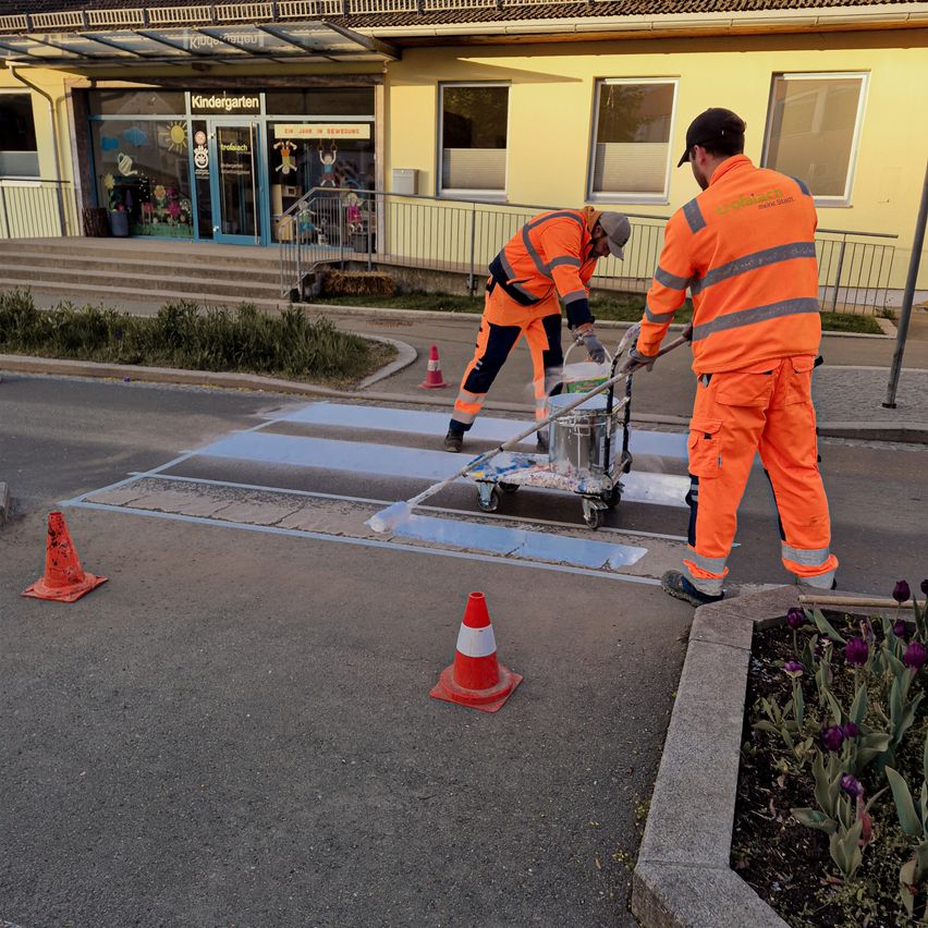 Two workers paint blue and white crosswalks in front of a yellow building with a shop named 'Kindergarten' and a sign for 'Tropflach'. The workers wear orange safety suits.