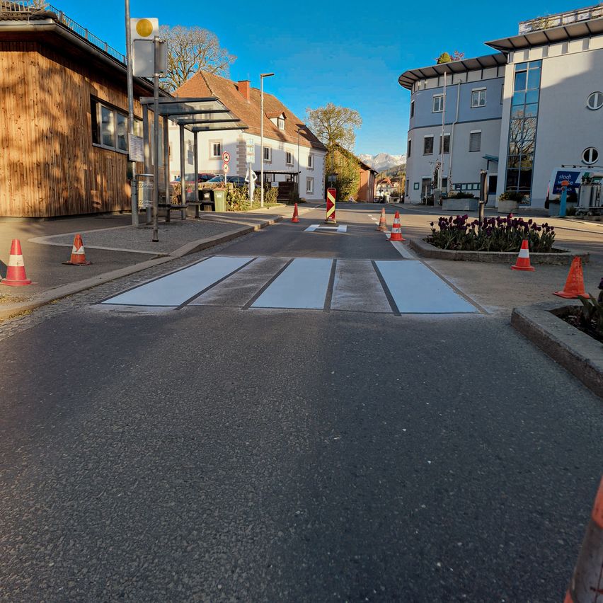 A road with a pedestrian crossing, marked with white lines and a zebra pattern, is surrounded by traffic cones. Buildings and trees are on either side.