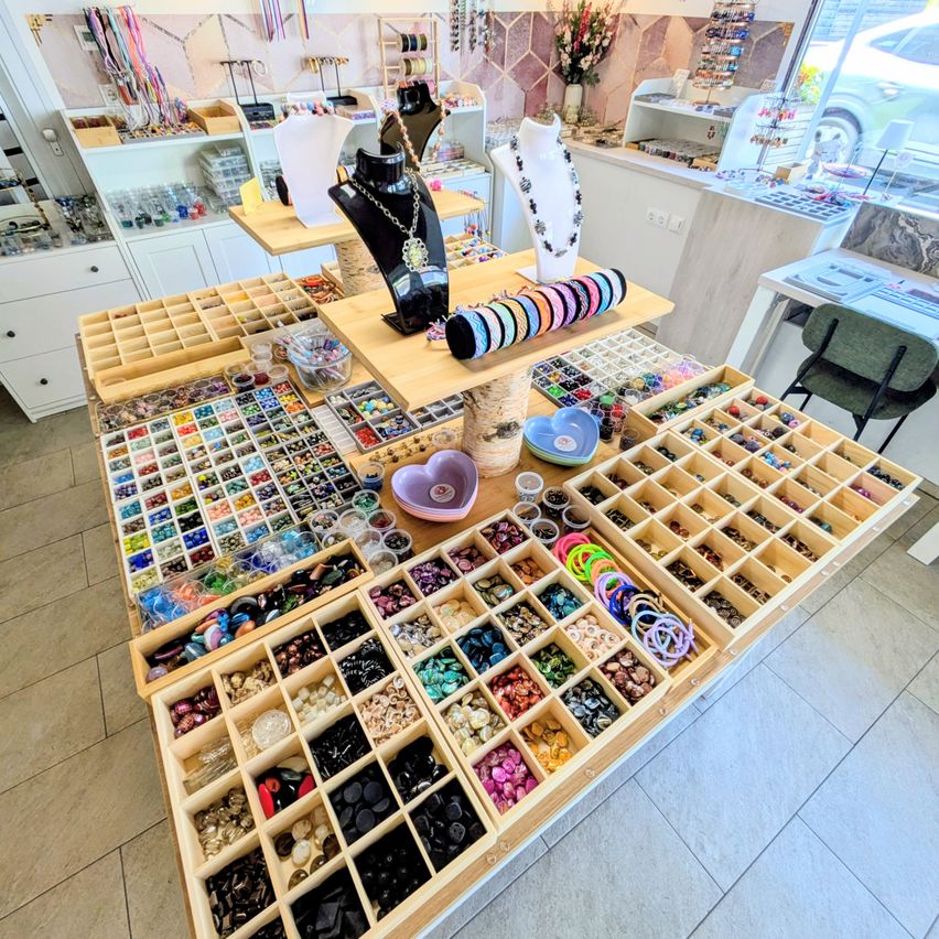 Ein Schmuckladen gefüllt mit verschiedenen Fächern, die Halsketten, Armbänder, Perlen und Knöpfe auf Holzregalen präsentieren. Auch Mannequins und eine Blumenvase sind vorhanden.