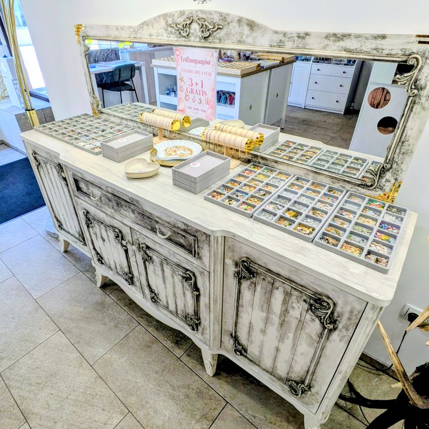 Eine elegant gestylte Schmuckvitrine mit verschiedenen Tabletts und Schachteln gefüllt mit Schmuck. Ein Spiegel reflektiert einen Raum mit Möbeln und einem Schild, das ein 3+1 gratis Angebot anbietet.