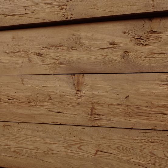 Eine Nahaufnahme von Holzbrettern, die natürliche Holzmaserungen, Äste und einige Abnutzungsspuren zeigt. Das Holz erscheint hellbraun mit sichtbarer Textur.