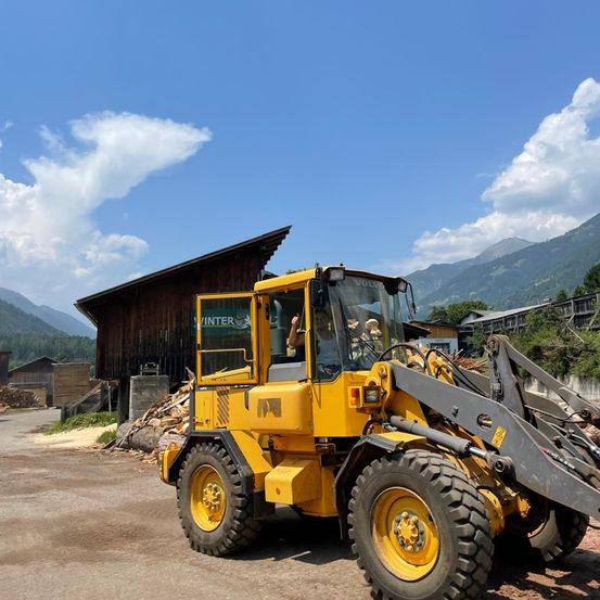 Ein gelber Baufahrzeug steht vor einer Scheune mit Bergen im Hintergrund.