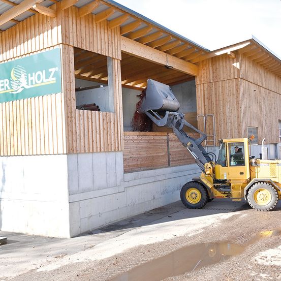 Ein gelber Gabelstapler lädt eine große Materialmenge in ein Holzgebäude mit dem Schild 'ER HOLZ'. Die Szene spielt sich draußen ab, mit einem nassen Boden und einem hellen Himmel.