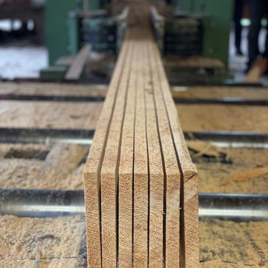 Eine Nahaufnahme von mehreren Holzbrettern, die auf einem Werkstatttisch mit Metalloberfläche gestapelt sind. Das Holz scheint frisch geschnitten und unbehandelt zu sein.