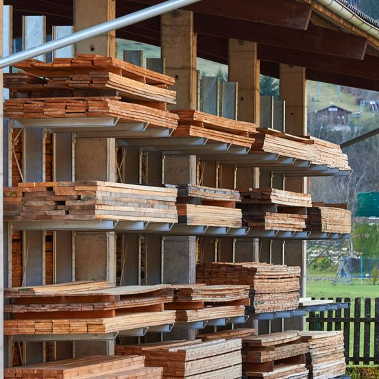 Eine Metall- und Betonlagerstruktur hält mehrere Schichten von Holzbrettern. Die Bretter sind ordentlich gestapelt, und die Struktur ist von einem Rasenfeld mit einem Zaun umgeben.