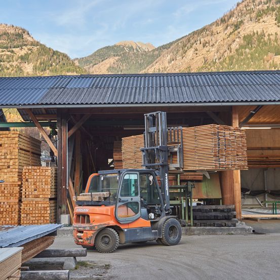 Ein Gabelstapler mit einer Holzladung steht in einem Lagerhaus mit Metalldach und Holzwänden, umgeben von Holzpaletten und Bergen im Hintergrund.